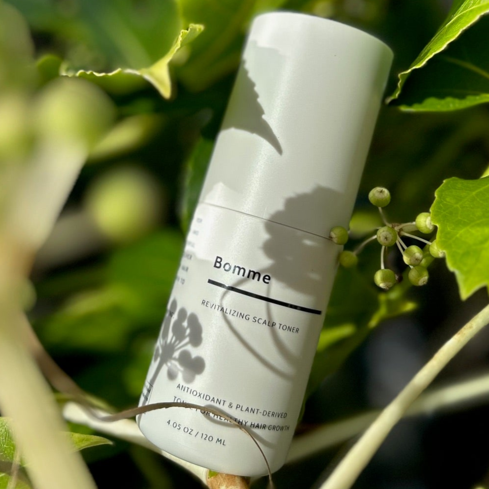 Bomme Revitalizing Scalp Toner placed among green leaves and buds. Natural setting highlighting plant-based formula.