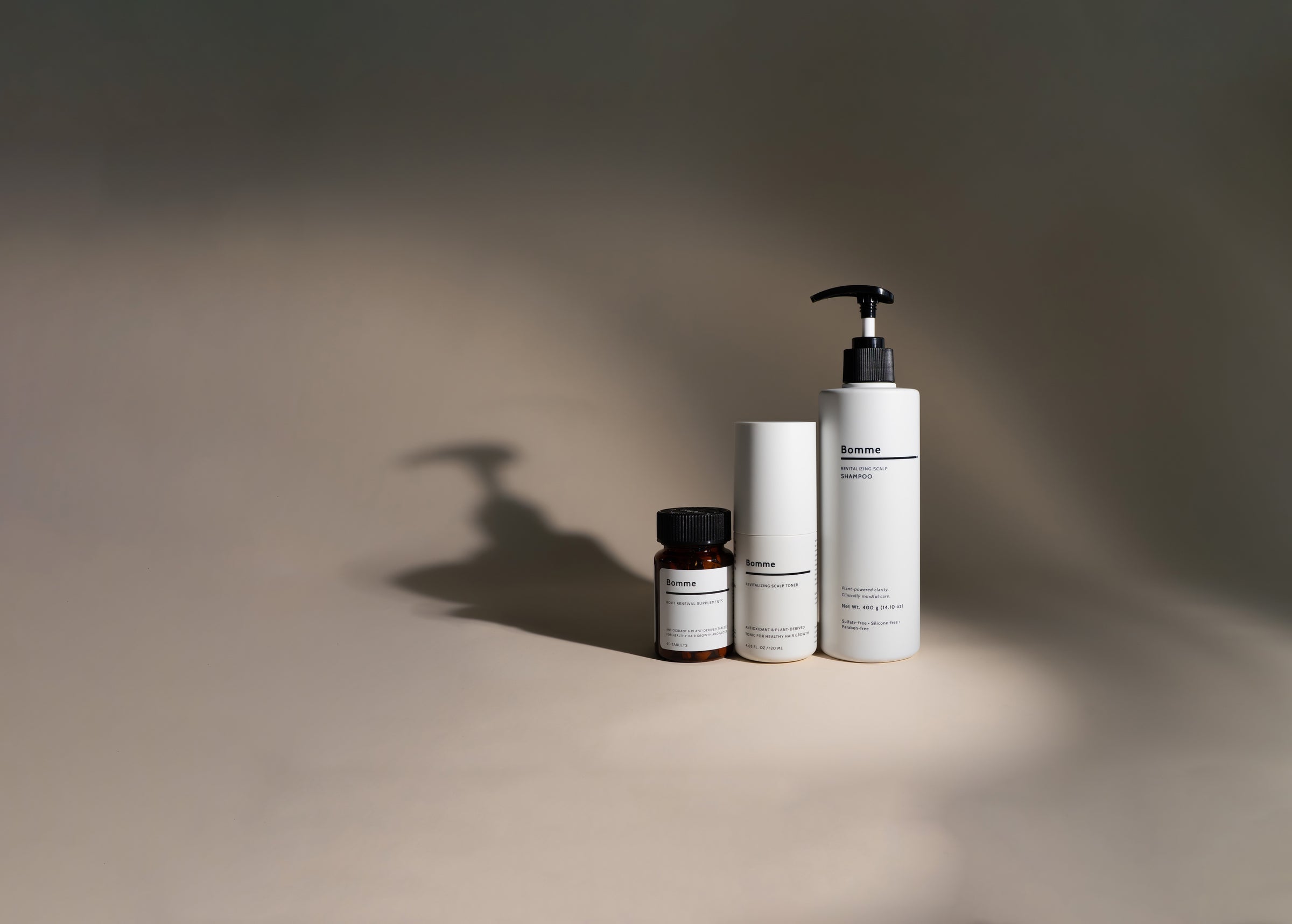 Bomme hair-care trio: Root Renewal Supplements, Revitalizing Scalp Toner, and Revitalizing Scalp Shampoo on a beige background with soft shadow.