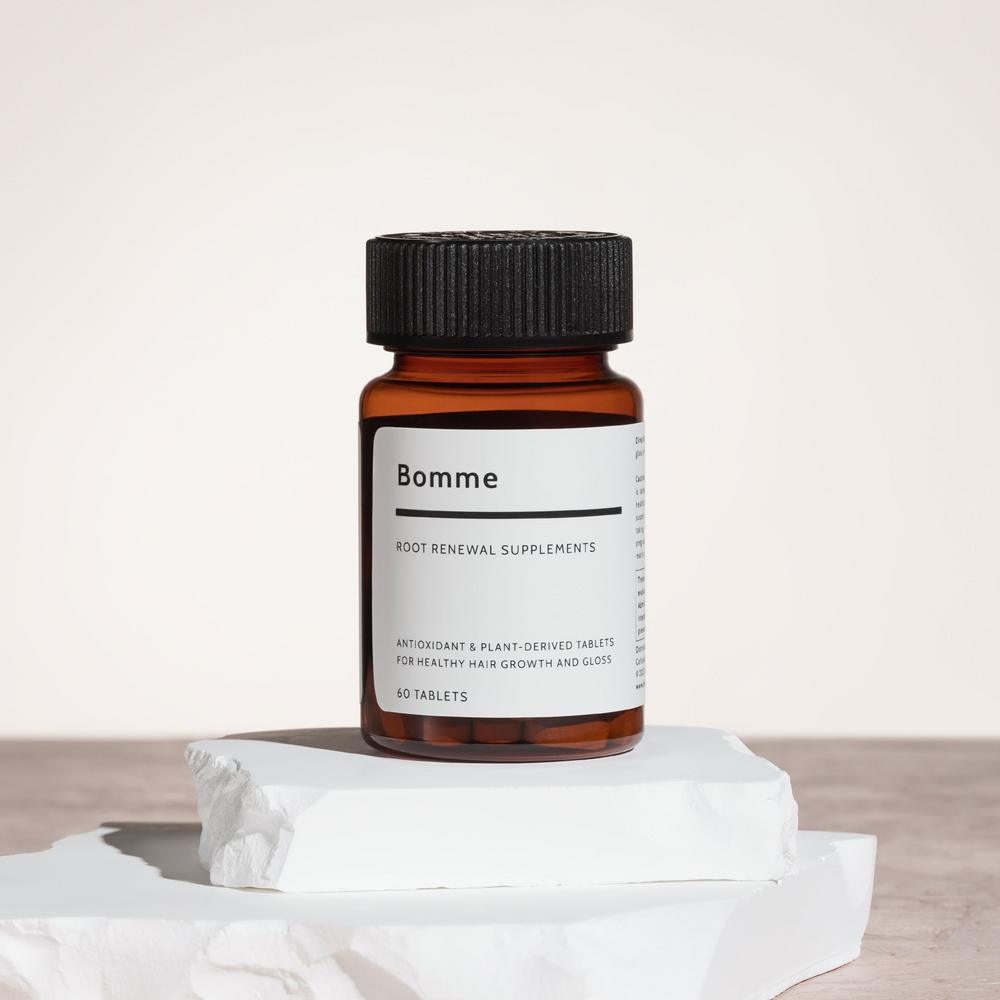 Amber bottle of Bomme Root Renewal Supplements with minimalist white label and black cap, displayed on white sculpted stone blocks against a neutral background.   