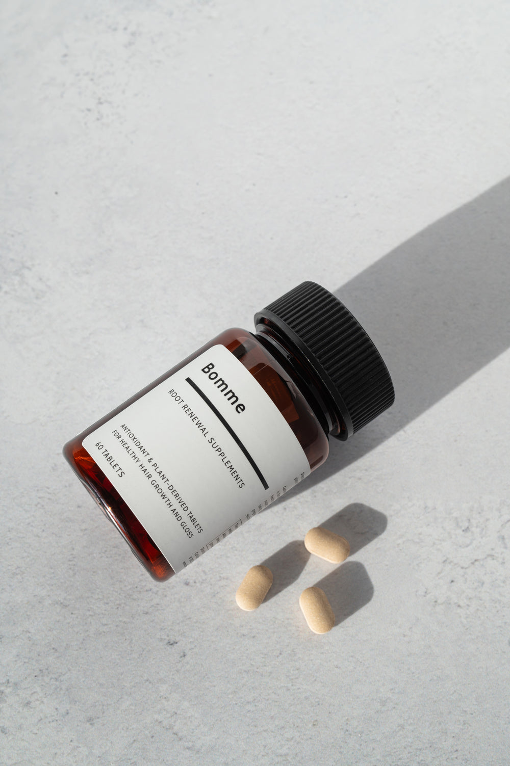 Bomme Root Renewal Supplements bottle with three beige tablets on a light stone surface. Plant-based antioxidant for healthy hair growth.