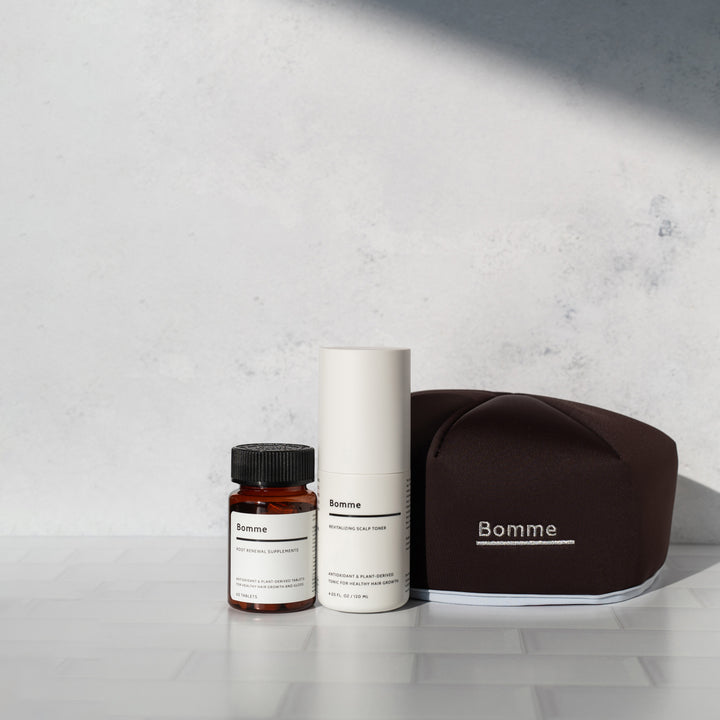 Bomme hair care set with supplements, scalp toner, and LED red light cap on a tiled surface with soft shadows