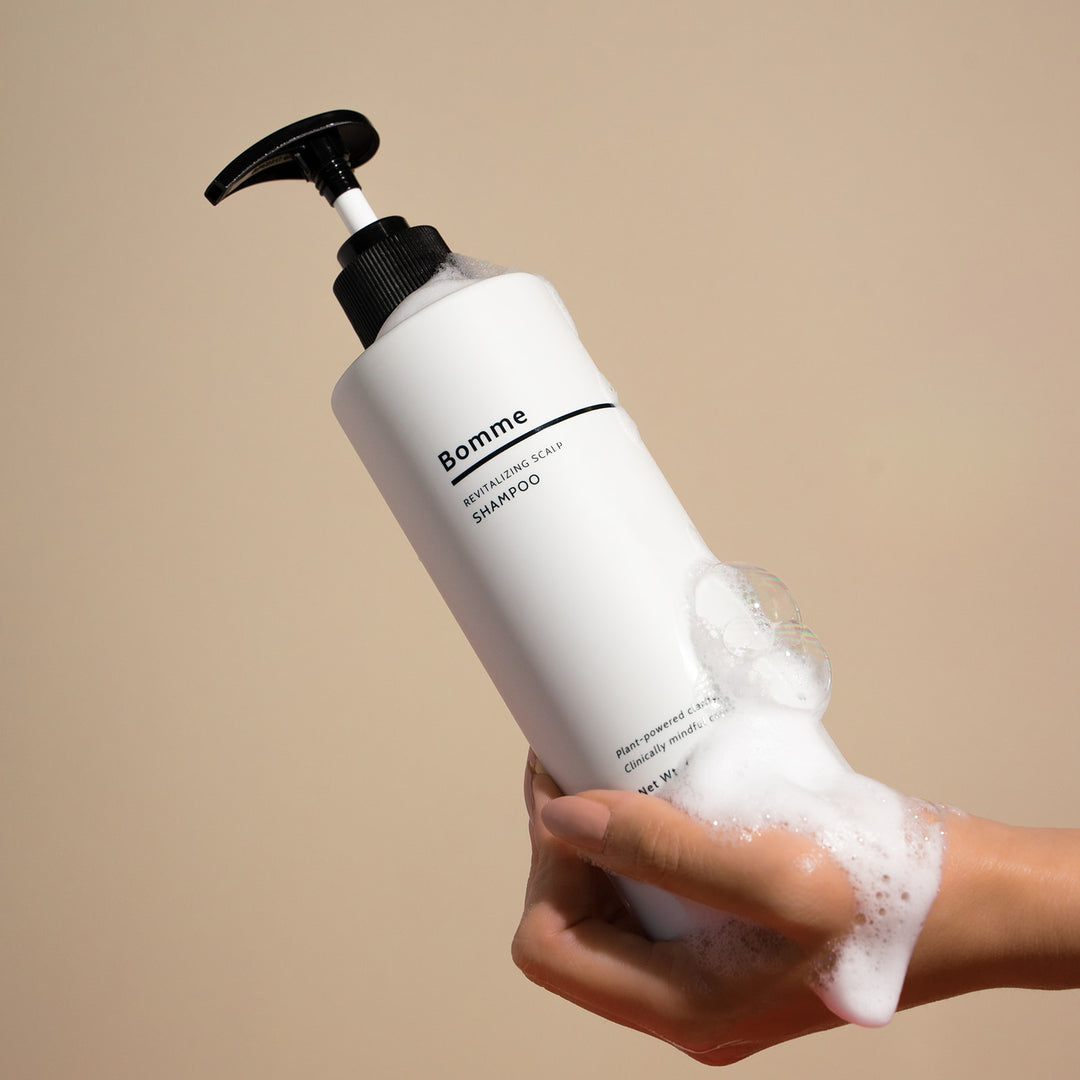 Bottle of Bomme shampoo held by a hand with soap suds on a beige background