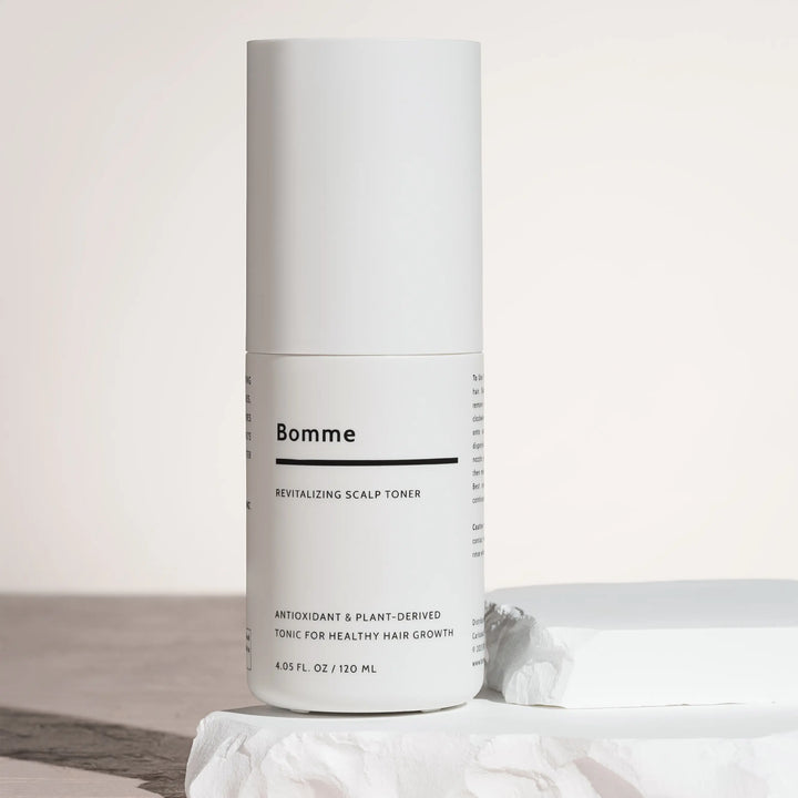 Bomme Revitalizing Scalp Toner bottle standing on white sculpted stone block with soft lighting. Antioxidant and plant-based toner for healthy hair growth.