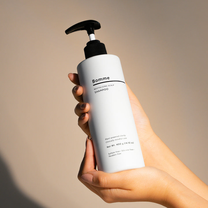 Hands holding a bottle of Bomme Revitalizing Scalp Shampoo with minimalist white packaging and pump dispenser.