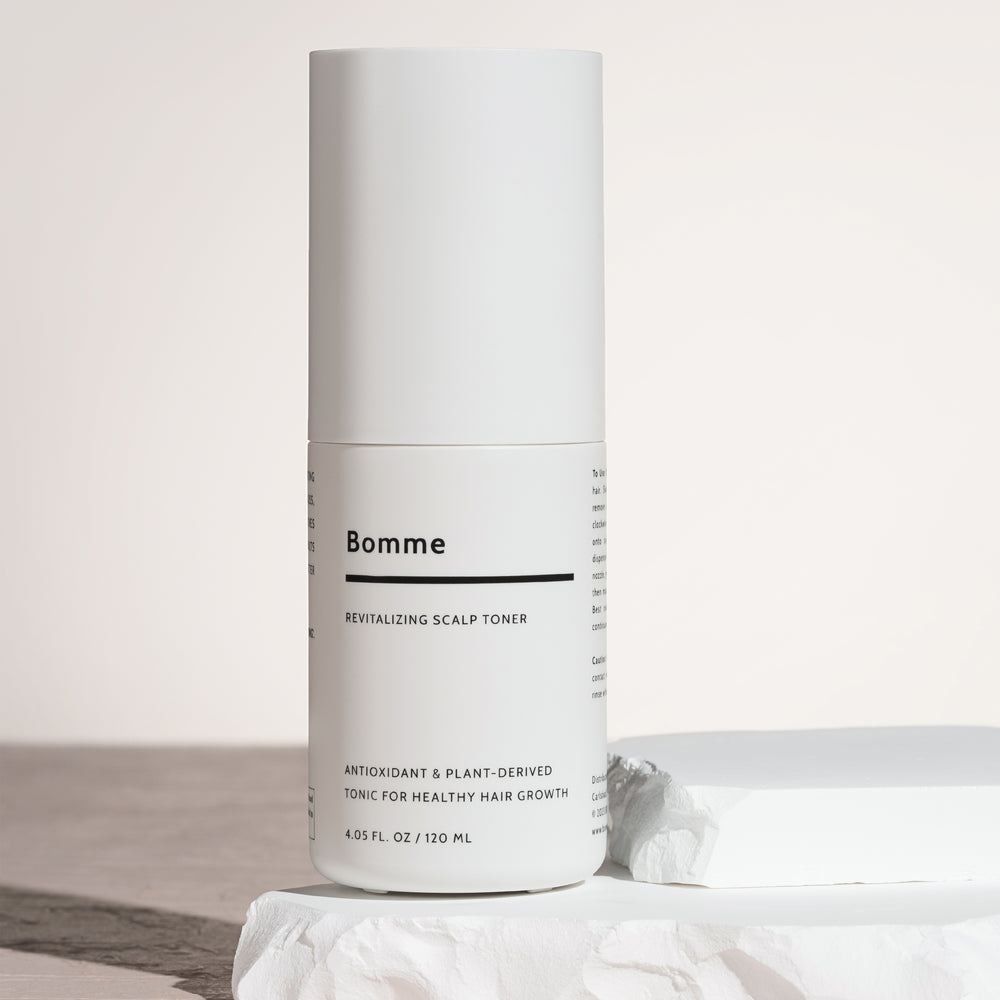 Bomme Revitalizing Scalp Toner bottle standing on white sculpted stone block with soft lighting. Antioxidant and plant-based toner for healthy hair growth.