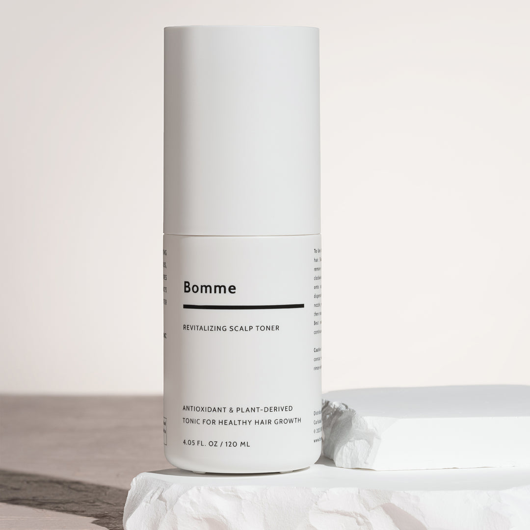 Bomme Revitalizing Scalp Toner bottle standing on white sculpted stone block with soft lighting. Antioxidant and plant-based toner for healthy hair growth.